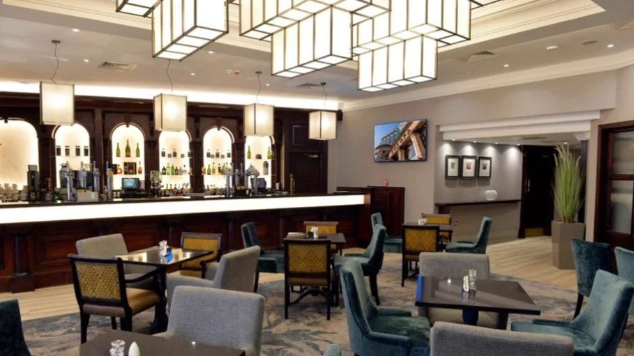 Modern bar and lounge area with a long white counter, bar stools, and a variety of bottles on illuminated shelves, with potted plants and framed pictures on the walls.
