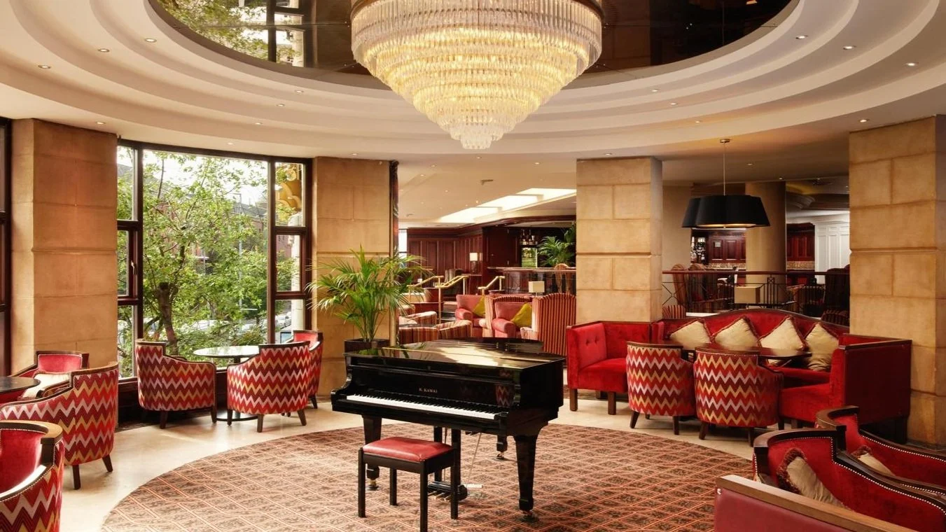 Elegant hotel lobby with a black grand piano at the center, surrounded by red and patterned chairs, a large chandelier hanging from the ceiling, large windows showing green trees outside, and warm lighting.