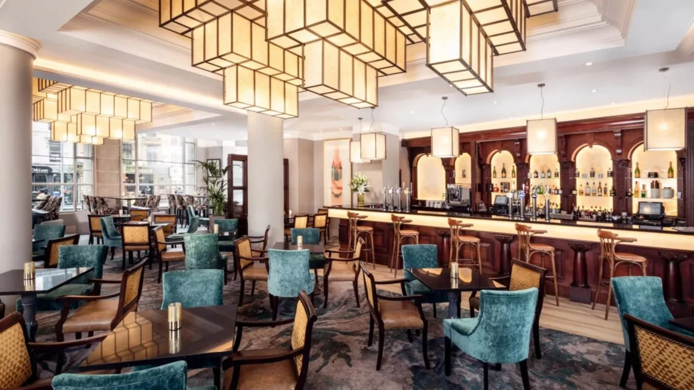 Interior of a modern bar and lounge with teal and beige chairs, dark tables, a long bar with wooden stools, illuminated shelves behind the bar with bottles, large pendant lights on the ceiling, and large windows letting in natural light.