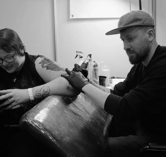 A man getting a tattoo while a woman is tattooing his arm in a tattoo studio.