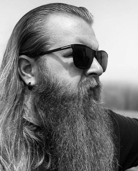 Close-up of a man with long hair and a full beard, wearing sunglasses and an earring, looking to the side in a black and white photo.