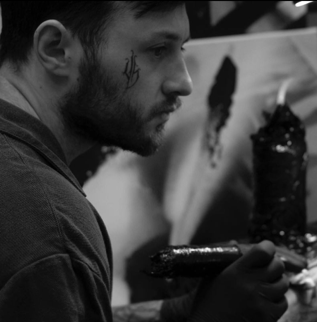 A young man with a tattoo on his cheek, working on a painting or sculpture in an art studio.