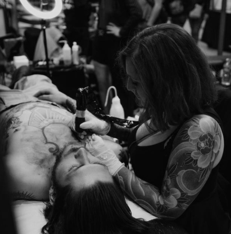 A woman with tattoos on her arm is tattooing a man's chest while the man is lying down on a tattoo artist's table. The setting appears to be a tattoo studio with people and equipment in the background.
