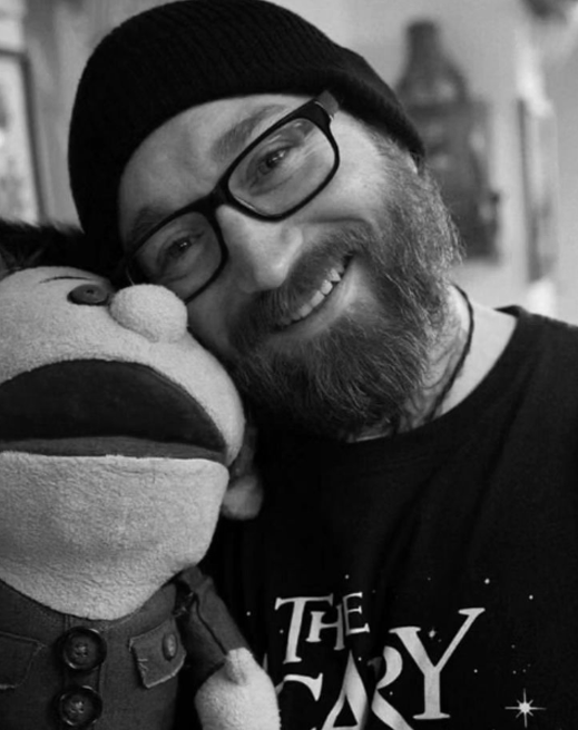 A man with glasses and a beard, smiling while posing with a stuffed puppet character.