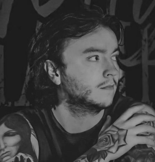 A black and white photo of a young man with dark, wavy hair and facial hair, looking to the right. He has tattoos on his arm, including a flower.