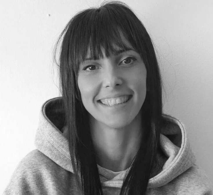 A woman with long dark hair and bangs smiling at the camera, wearing a hoodie.