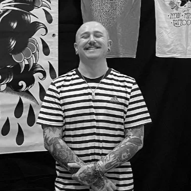 A smiling man with tattoos on his arms, wearing a striped t-shirt, standing in front of a tattoo shop display, with artwork and T-shirts hanging in the background.