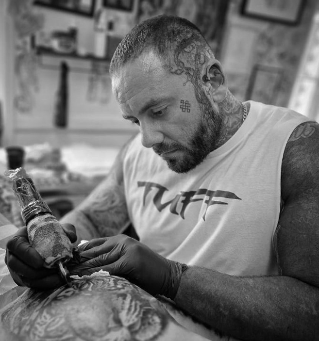 A man with tattoos, wearing a T-shirt, is focused on applying a tattoo to another person's leg, wearing black gloves, with a detailed tattoo design.