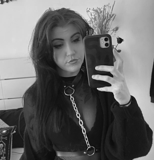 Black and white photo of a young woman taking a mirror selfie with her phone. She has long dark hair, makeup, and a choker necklace with a chain. She is wearing a dark top and standing in a room with a plant and a pillow in the background.