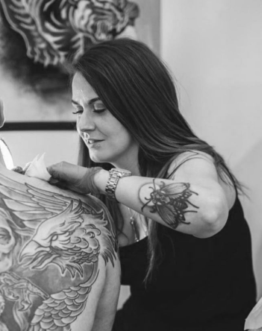 Woman with long dark hair tattooing a man's chest, featuring a dragon design; woman has a butterfly tattoo on her arm and is focused on tattooing.