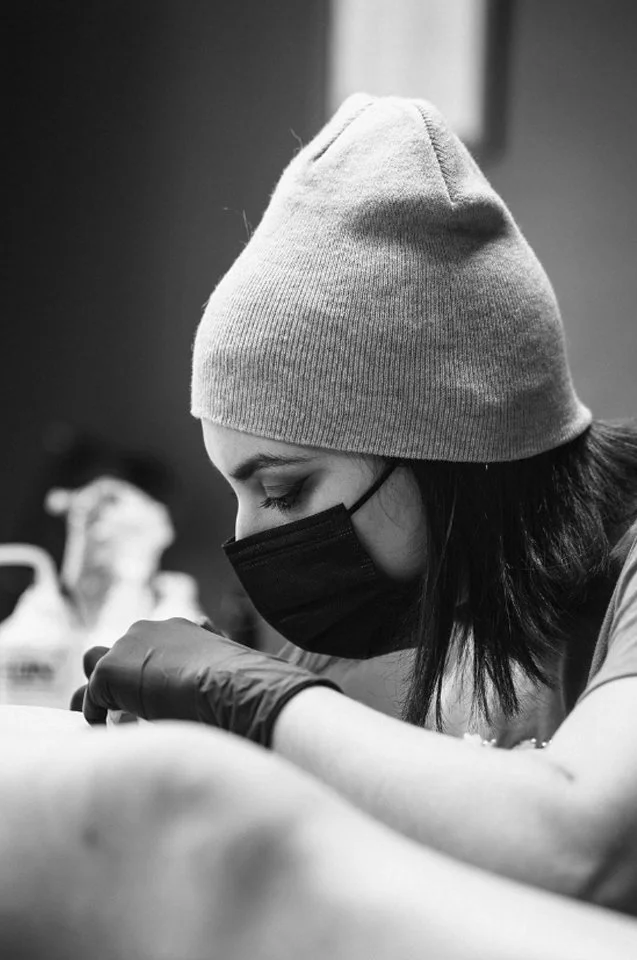 A woman wearing a beanie, face mask, and gloves, performing a tattoo or medical procedure.