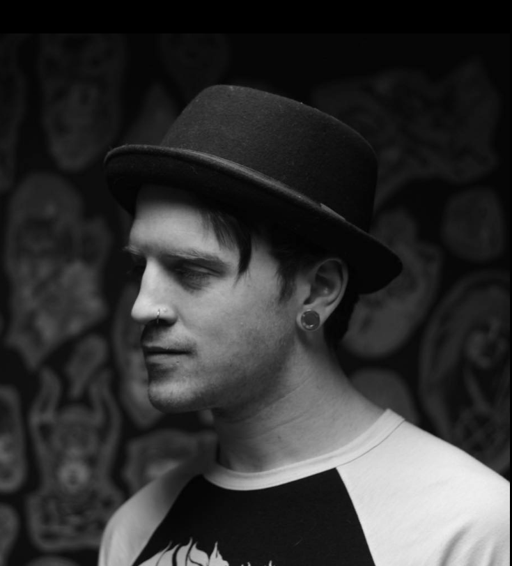 Black and white photo of a young man wearing a dark hat, with facial piercings and earrings, and a casual shirt, standing in front of a tattooed wall.