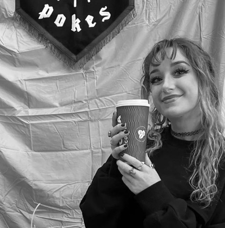Young woman with curly hair and choker necklace holding a drink cup, smiling, in front of a backdrop with a sign that says 'POKE'S' in black and white.