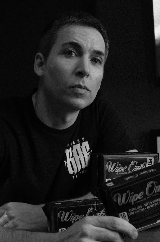 A person with short hair sitting at a table in a dark room, holding packs of Wipe Off cleaning wipes, looking at the camera.