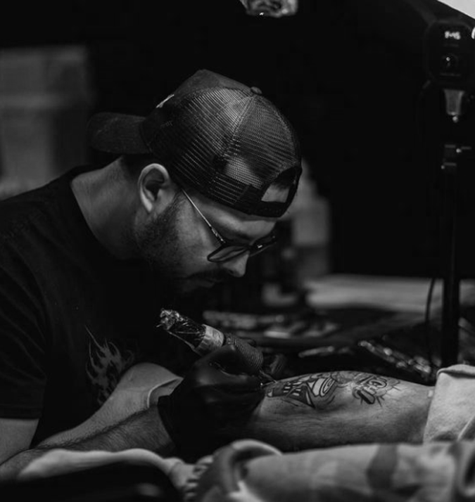 A man with glasses and a cap working on a tattoo tattooing on someone's arm in a studio.