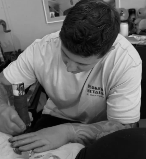 A man with short dark hair, wearing a white t-shirt, is focusing on a task with his right hand resting on another person's arm. The background shows kitchen shelves with jars and bottles.
