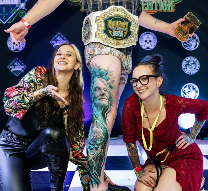 Two women crouching and smiling beside a person with a championship belt, standing in the center. The person in the middle has a large, detailed tattoo of an elderly woman's face on one leg, and is holding a trophy and a card. The background features a dark backdrop with various logos.