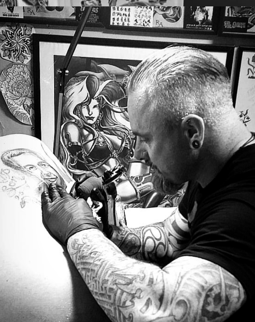 Tattoo artist working on a client's arm, surrounded by framed artwork in the background.