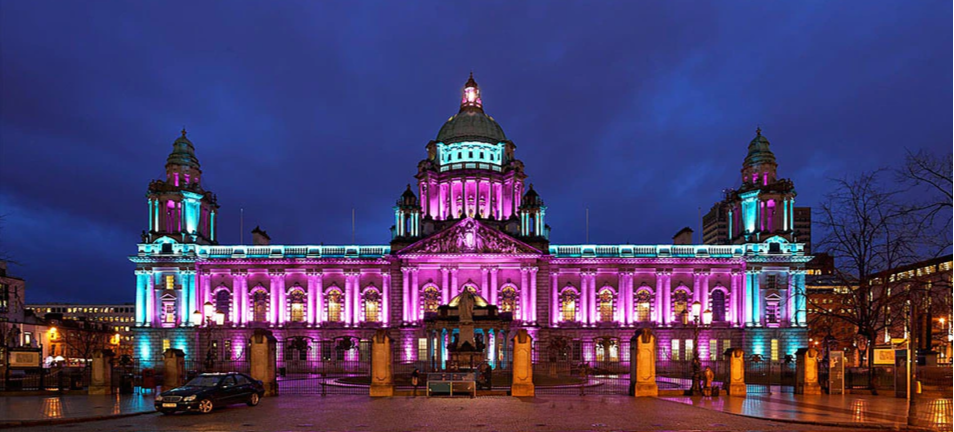Historic building illuminated with pink, purple, and blue lights at night, with a cloudy sky in the background.