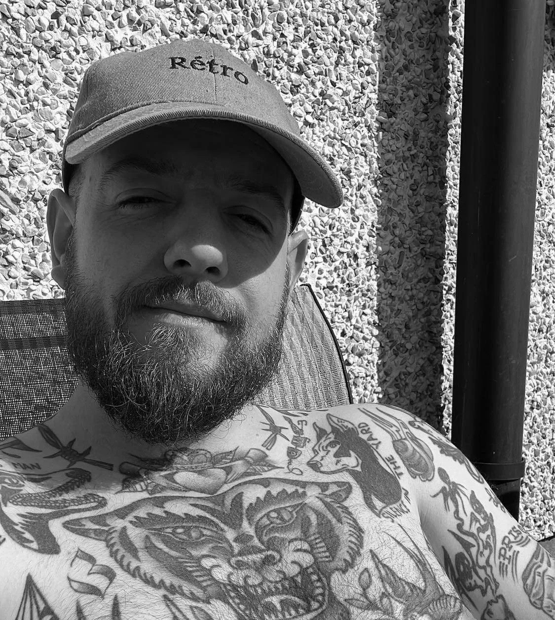 A man with a beard wearing a cap and showing his tattoos on his chest and arms, sitting outside.