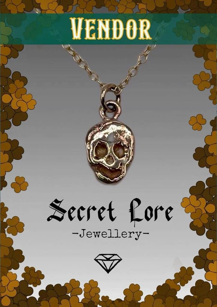 A jewelry necklace with a skull pendant hanging from a chain, displayed against a packaging with floral decorations and labels 'VENDOR' and 'Secret Lore Jewellery'.