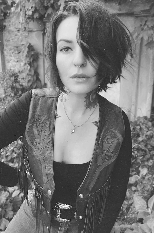 A young woman with short, tousled dark hair, wearing a leather vest with fringe details, a black top, and a necklace with a star-shaped pendant. She has a nose ring and is standing outdoors with foliage and a wooden fence in the background.