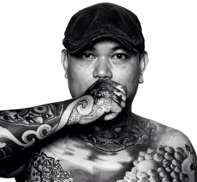 A black and white photo of a shirtless man with extensive tattoos covering his chest and arm. He is wearing a cap and has his hand raised to his face, covering his mouth.