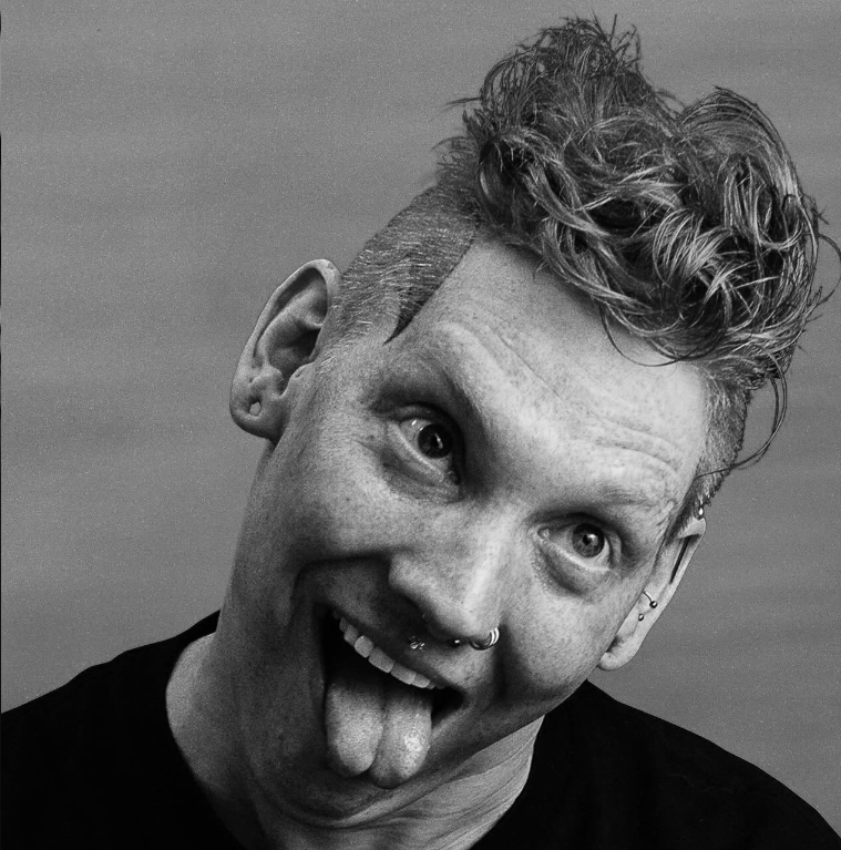 A Black and white photo of a person with short curly hair sticking out their tongue and looking upward, with multiple facial piercings.