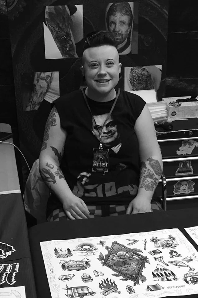 A person with short dark hair smiling, wearing a necklace with a badge labeled 'Artist,' seated at a table with artwork and tattoo sketches, with tattoos on arms, in a room decorated with various drawings and paintings.