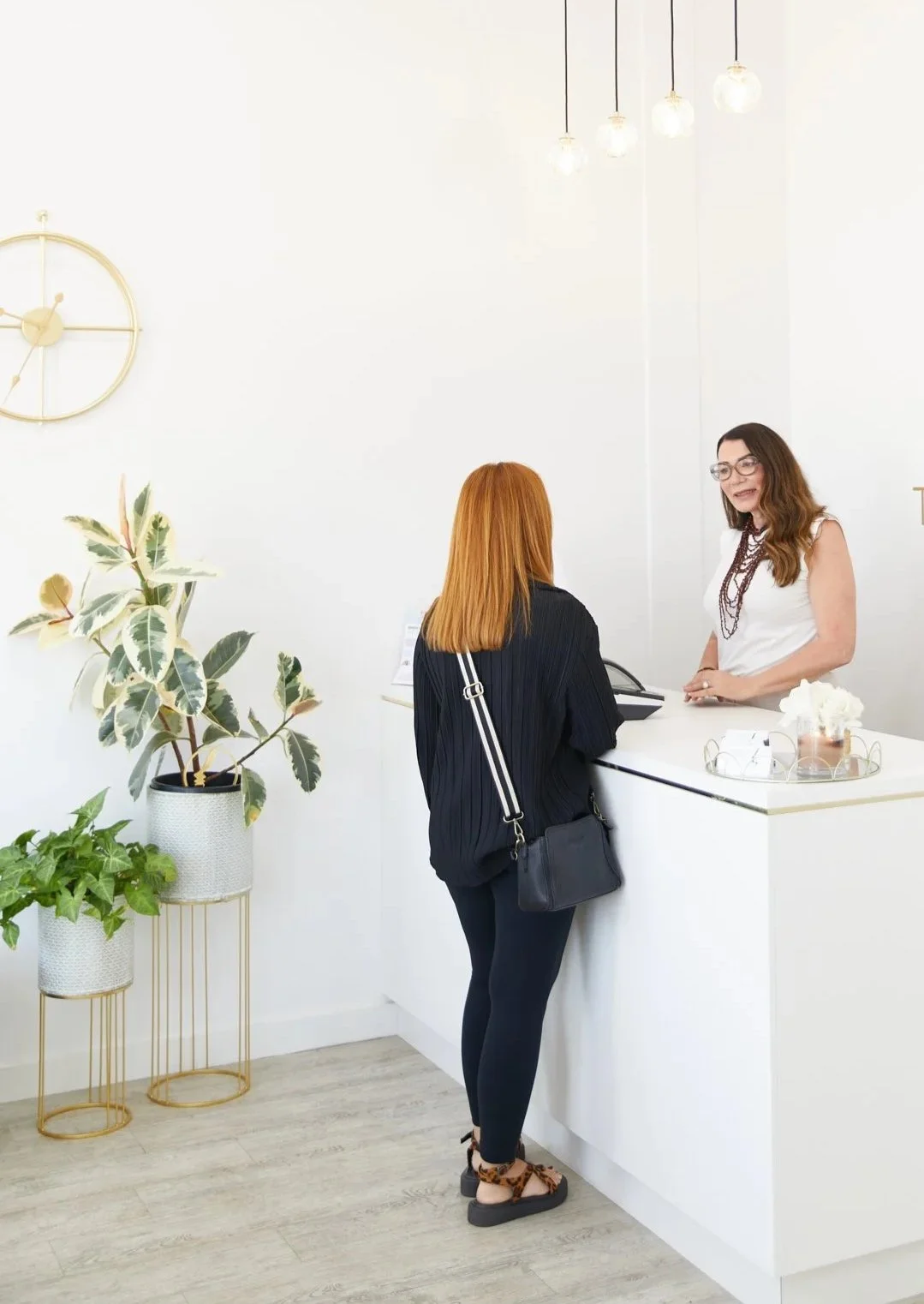A woman with red hair checks in at a reception desk while a receptionist with brown hair and glasses interacts with her in a modern, brightly lit office or hotel lobby.