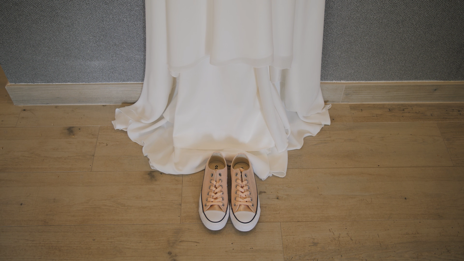 Sapatos branco de lona ao lado de um vestido branco com o chão de madeira ao fundo.