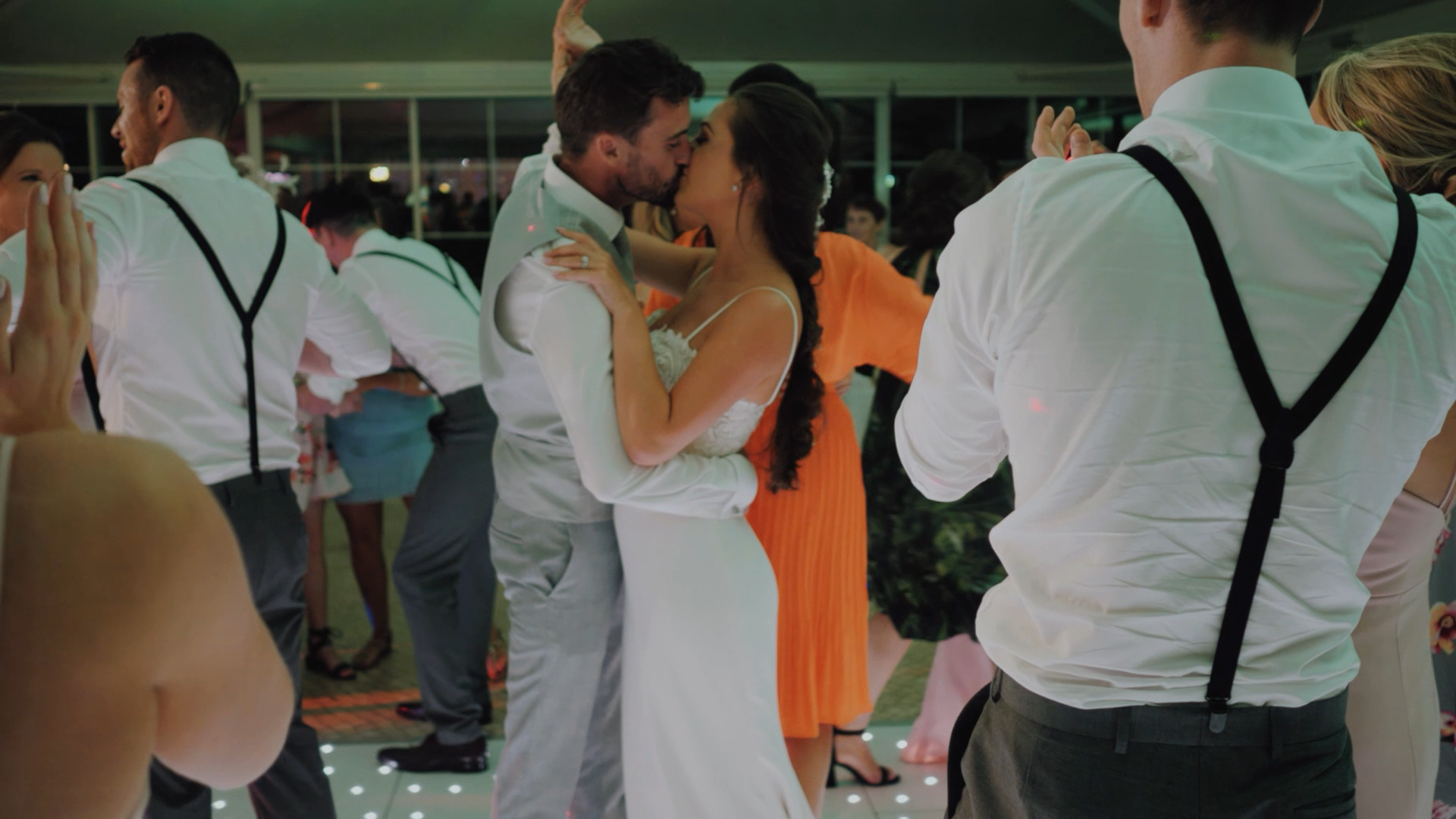 Casal de noivos se beijando na pista de dança em casamento, cercado por convidados dançando e celebrando.