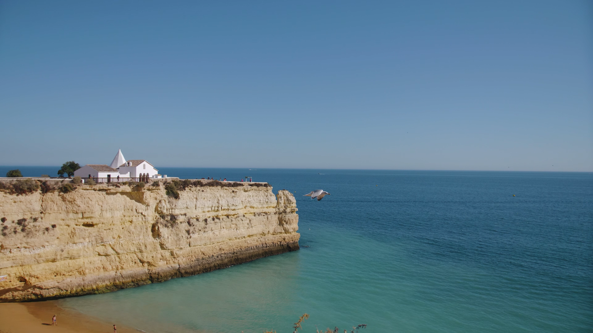 Cena de uma falésia com uma igreja branca ao fundo, o mar azul à direita e uma praia com algumas pessoas na areia na parte inferior da imagem, durante um dia ensolarado.