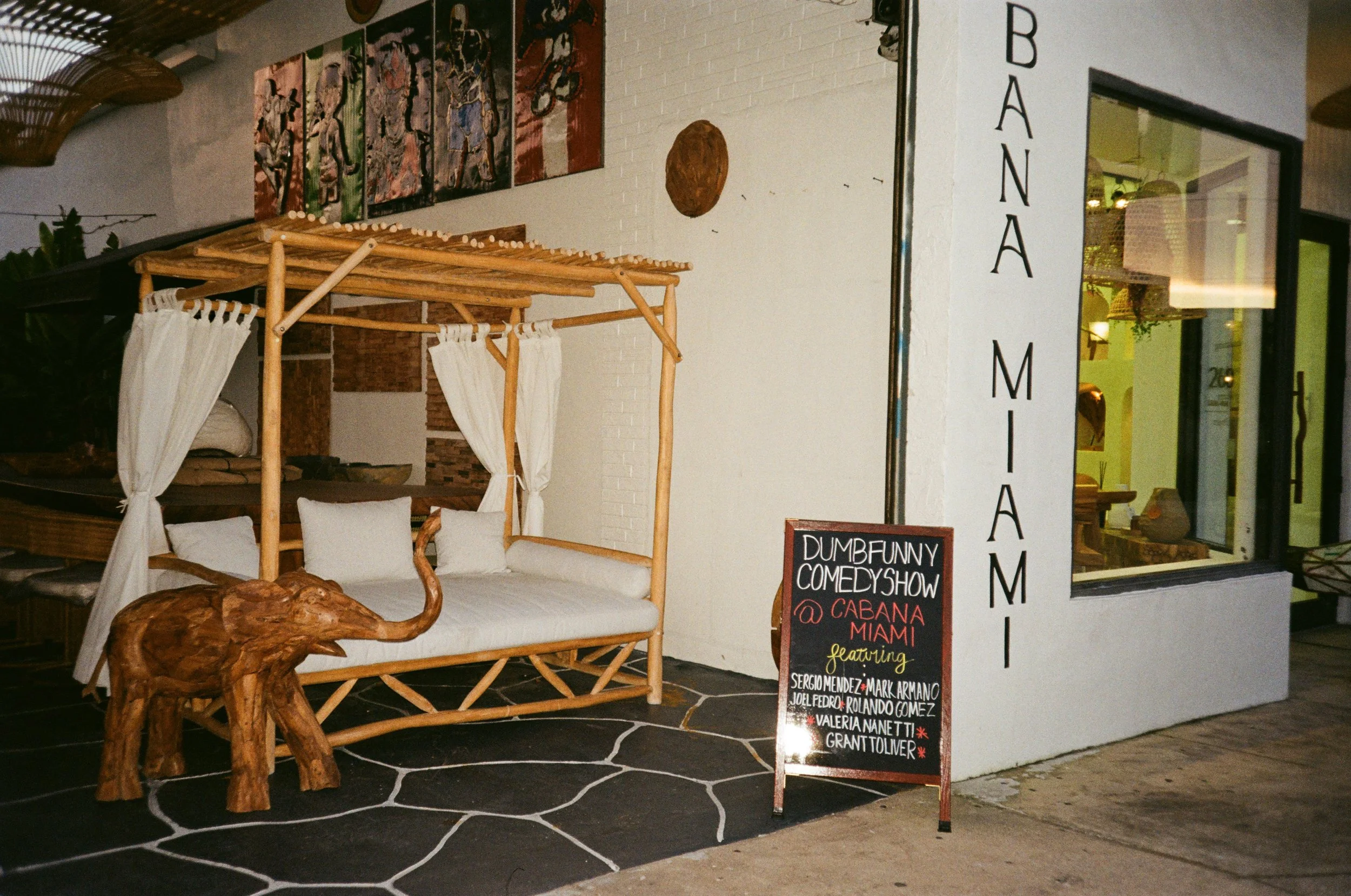 Entrance of a Miami-based venue called Cabana, decorated with a canopy bed and a wooden elephant sculpture in front. A chalkboard sign advertises a comedy show featuring several performers.