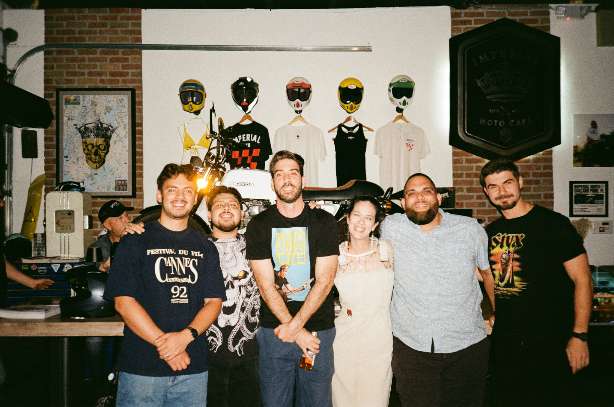 Group of six people smiling and posing in a bar or cafe with motorcycle-themed decor, including helmets, shirts, and a motorcycle, in the background.