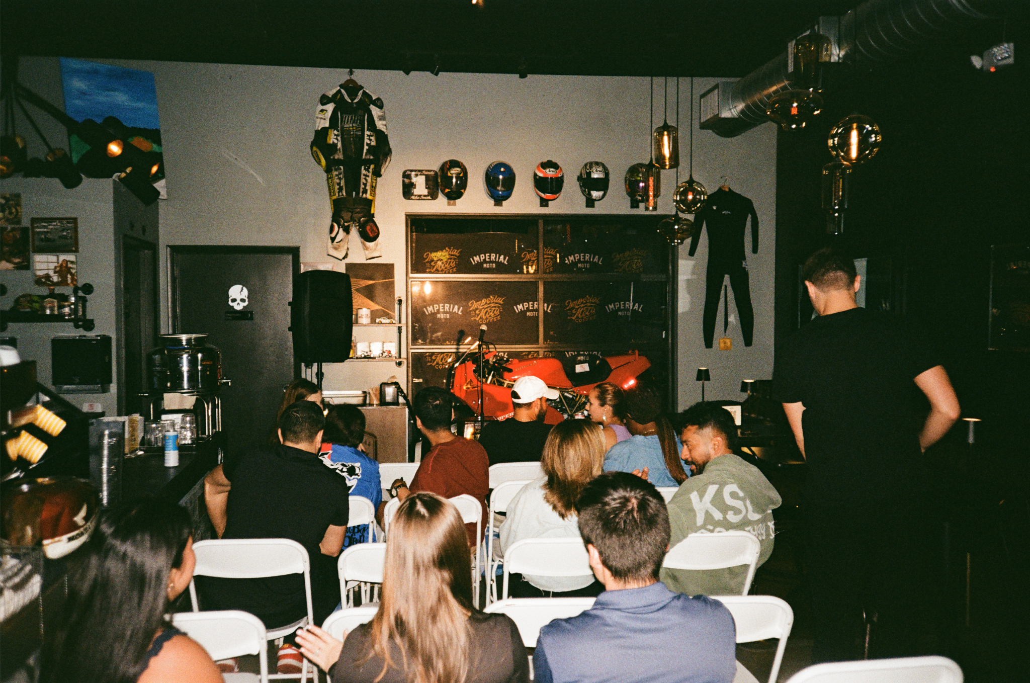 An indoor gathering with people seated in white chairs watching a stage with motorcycles, helmets, racing suits, and promotional banners.