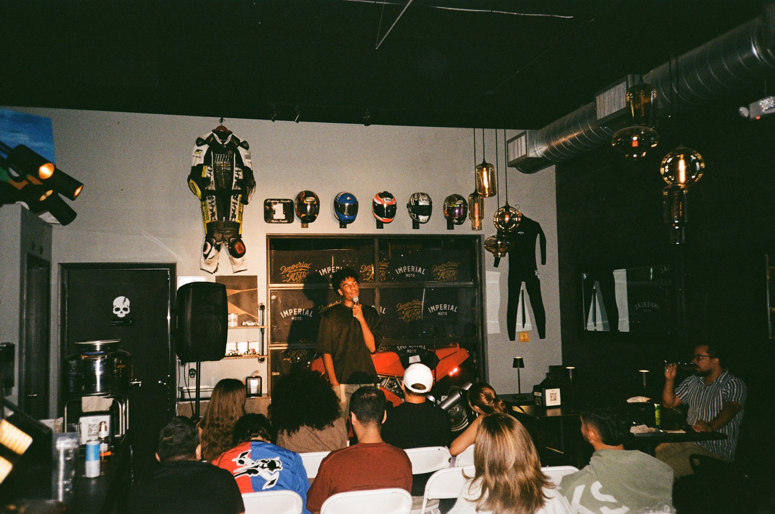 A man with curly hair speaking into a microphone at a motorcycle-themed comedy show or event, with audience seated in front. The background includes motorcycle helmets, a racing suit, a motorcycle, and motorcycle-related memorabilia, with decorative 