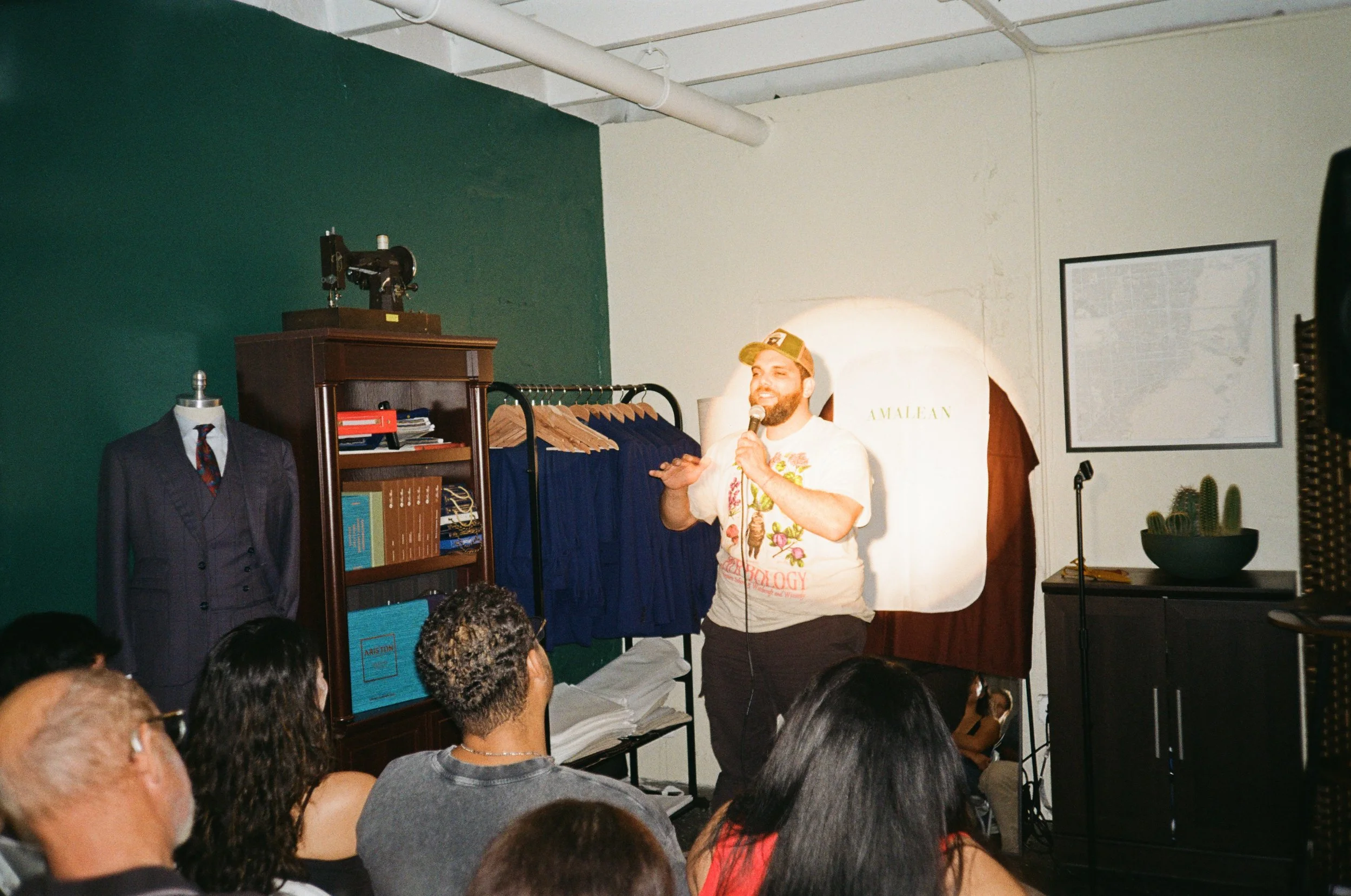 A man with a beard and a tan baseball cap speaking into a microphone in front of an audience in an indoor space with clothing and vintage items.