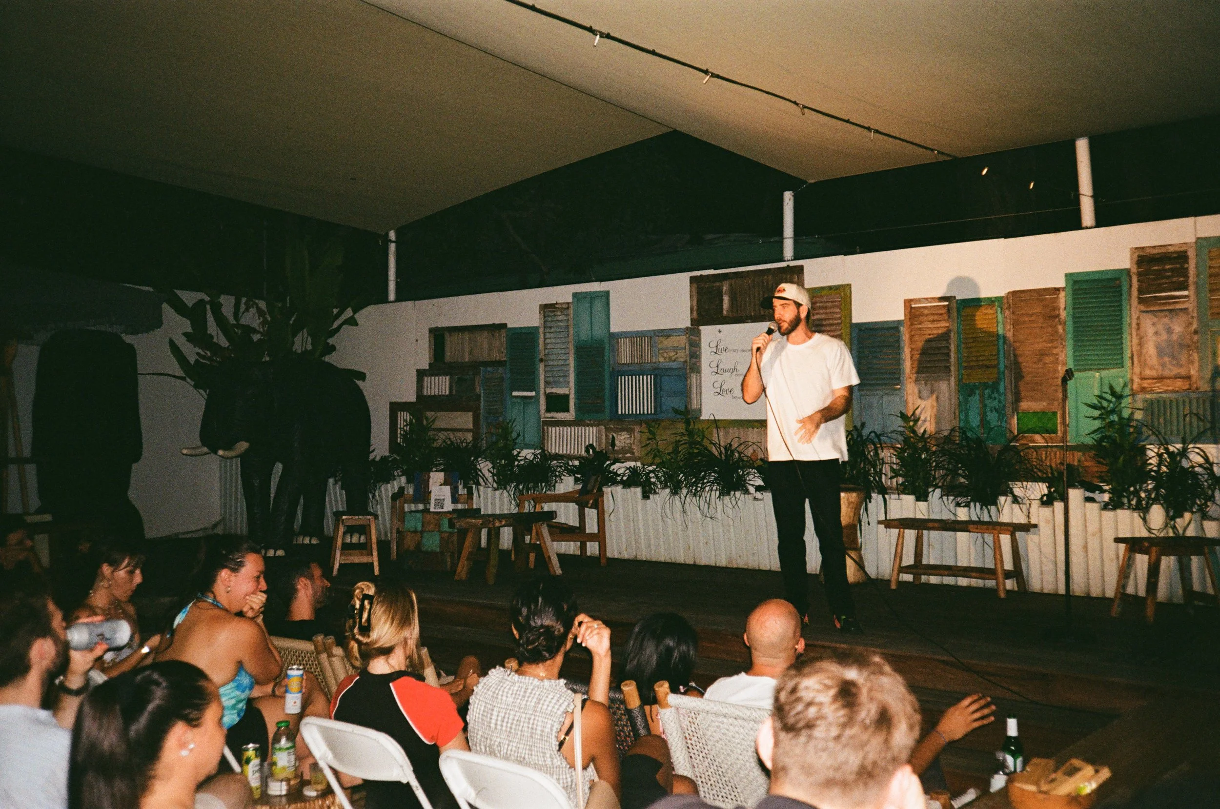 A man stands on a stage holding a microphone, performing in front of an audience. The stage background features colorful, distressed window shutters and potted plants.