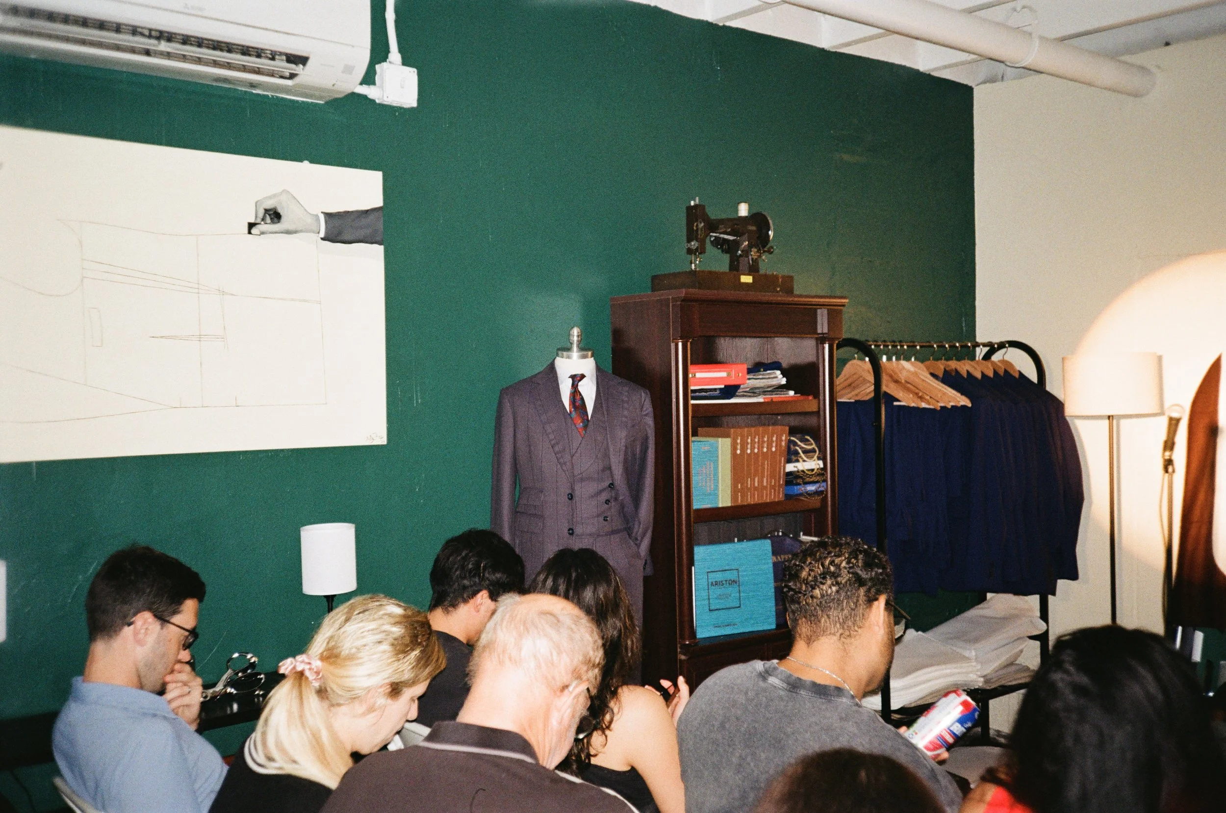 A group of people sitting inside a room with a green wall. There is a mannequin dressed in a suit, shelves with books and folders, a clothing rack with blue garments, a lamp, and a drawing on the wall.