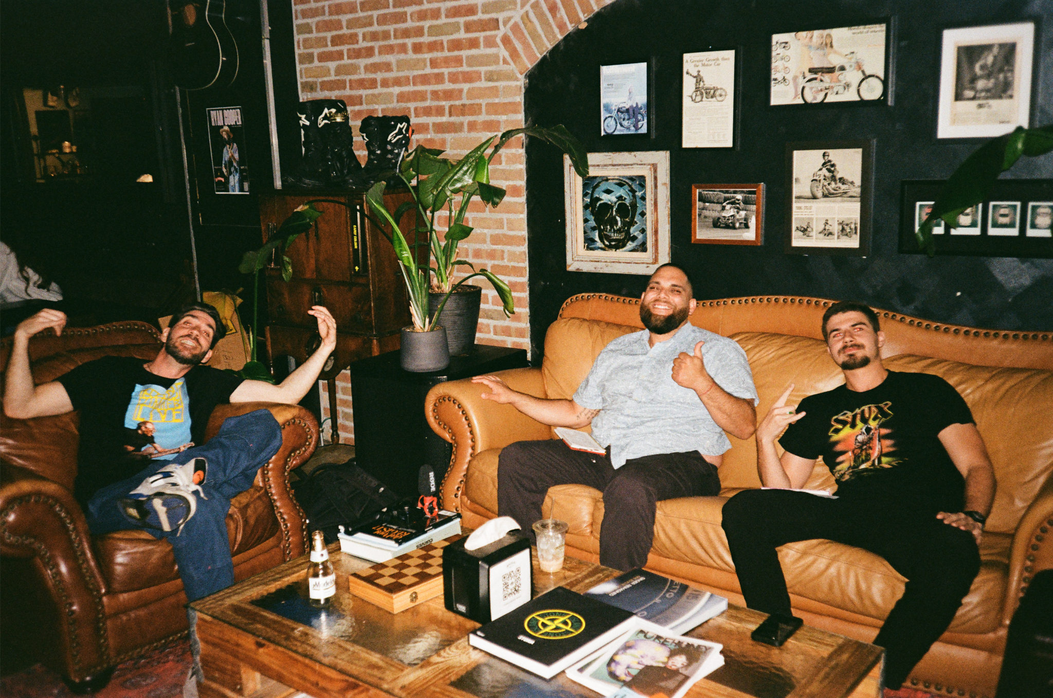 Three men sitting on leather couches in a cozy, decorated room, smiling and making relaxed gestures. The room has a brick wall, framed artwork, a large green plant, and a coffee table with books, a tissue box, a chessboard, a drink, and a bottle.