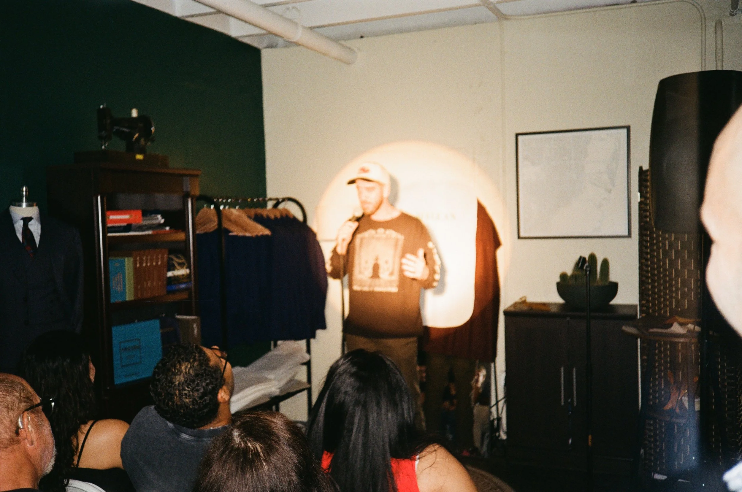A man wearing a cap and black sweatshirt speaking into a microphone on a small stage, with an audience seated in front of him, indoor setting with clothes on racks and a mannequin.