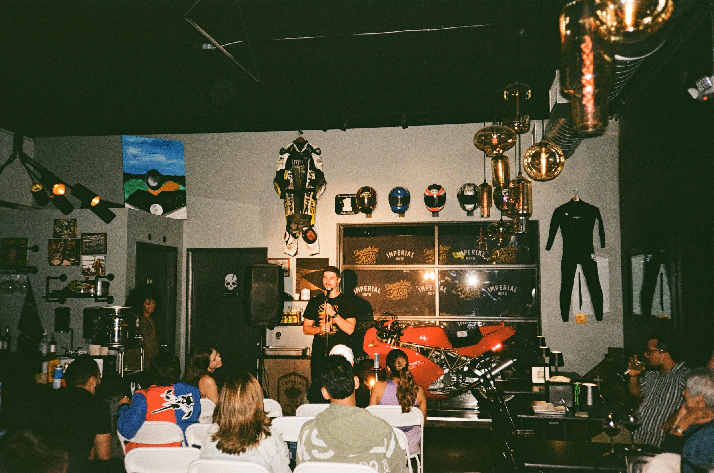 People attending a comedy or stand-up show in a venue decorated with motorcycle gear, helmets, and racing suits, with a red motorcycle on stage and an audience seated facing a performer.