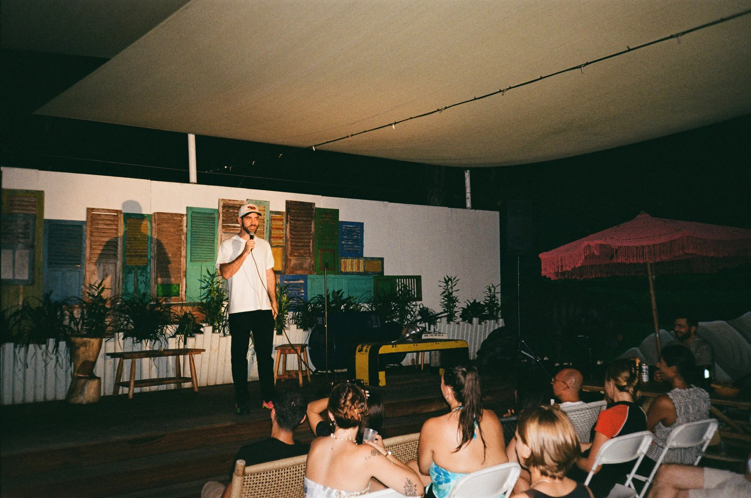 A man in a white T-shirt, cap, and black pants stands on a small stage holding a microphone, performing in front of an audience at an outdoor venue. The background features colorful window shutters, potted plants, and a pink umbrella to the right. Au