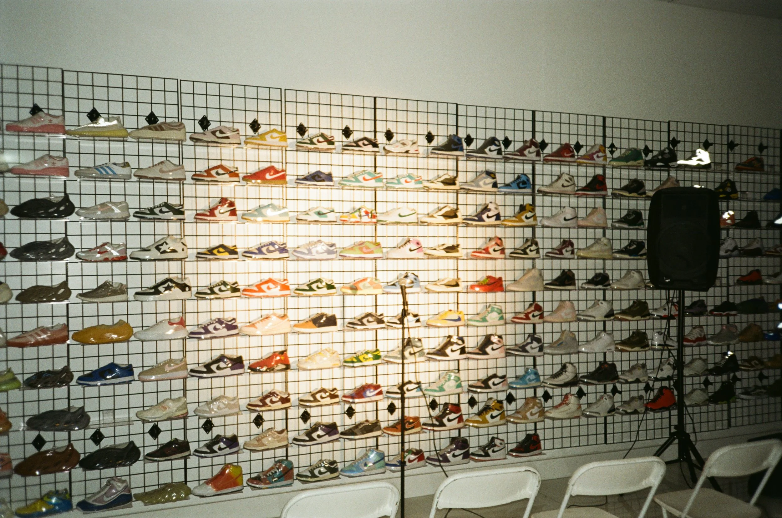 Display of various sneakers on black wire grid wall at a sneaker store or event, with a speaker and chairs in front.