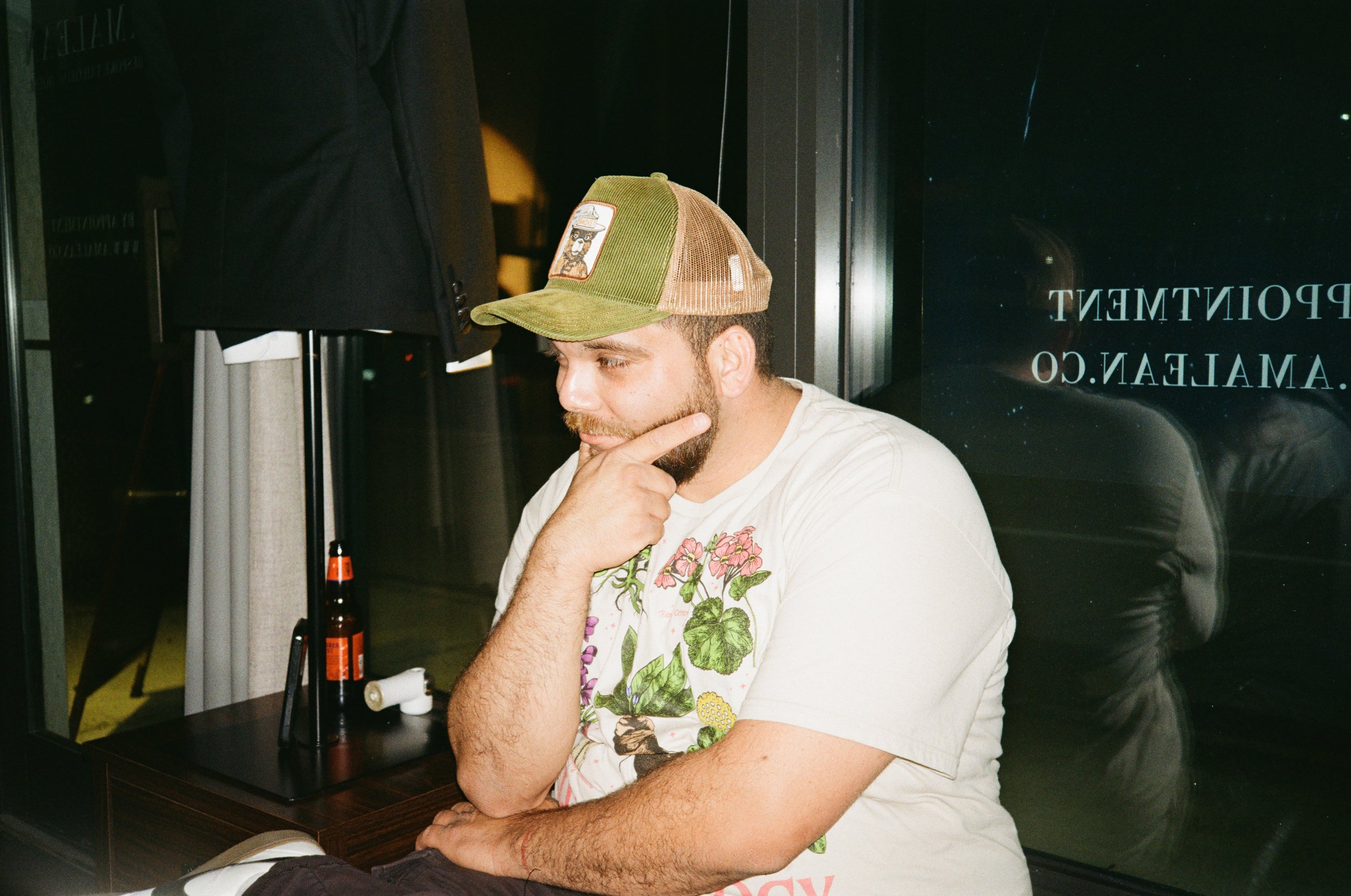 A man in a colorful t-shirt and a trucker hat sits at a table with his hand on his chin, appearing to be deep in thought. There are a couple of bottles on the table and a beer bottle placed upright behind them.