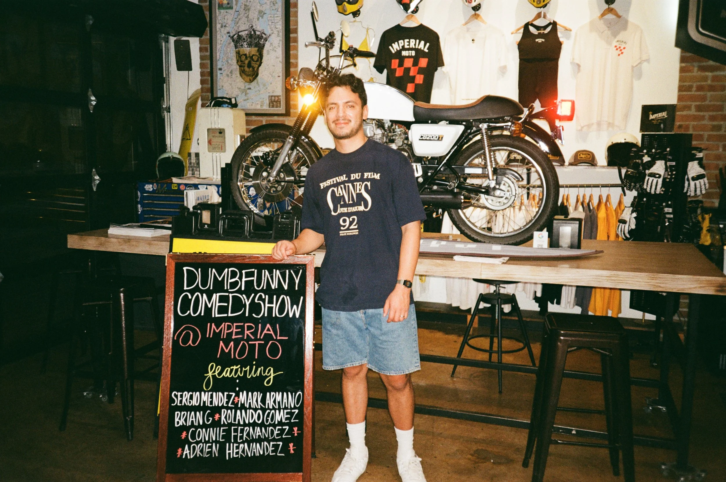 A young man standing next to a chalkboard sign promoting a comedy show at Imperial Moto, featuring performers Sergio Mendez, Mark Armano, Briang, Rolando Gomez, Connie Fernandez, and Adrien Hernandez. he is in an indoor space decorated with clothing,