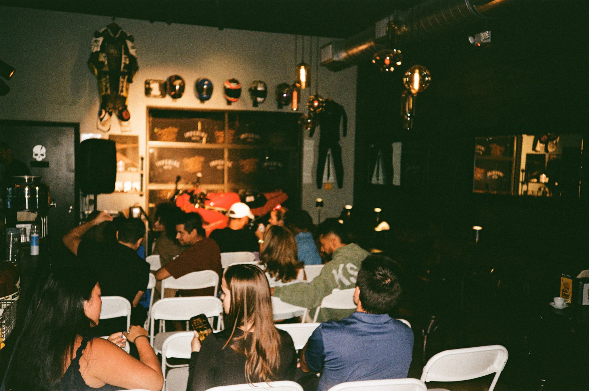 People sitting on white chairs watching a motorcycle racing event in a dimly lit indoor venue, with motorcycle helmets and racing memorabilia on the wall.