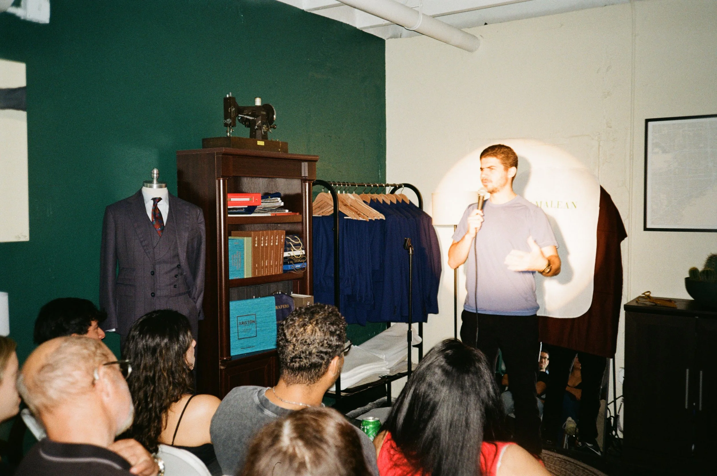 A man holds a microphone and speaks to an audience in a room with a clothing rack, a bookshelf, and a mannequin dressed in a suit.