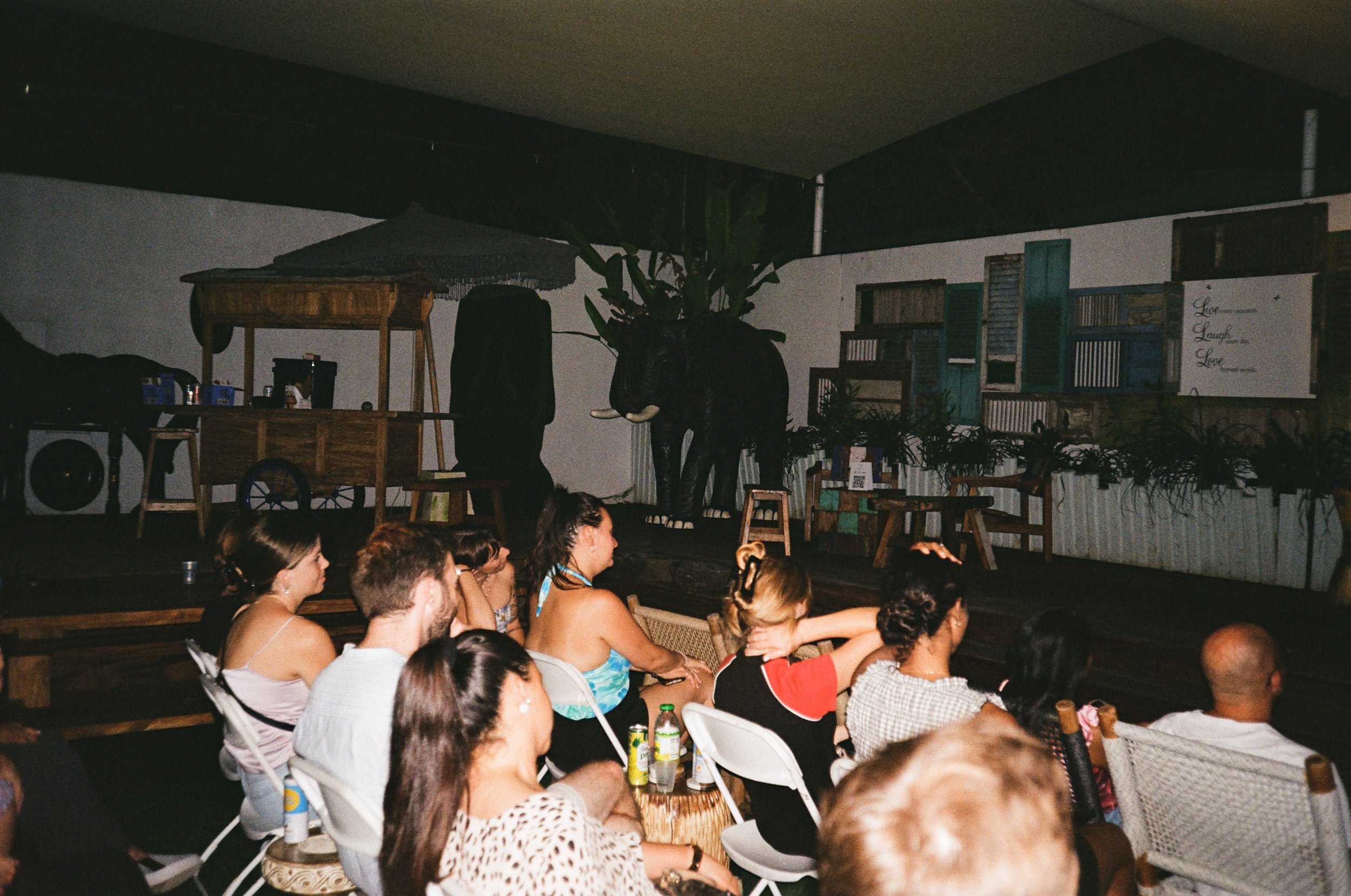 Audience watching a performance on a small stage with a backdrop of vintage shutters and a painted elephant.